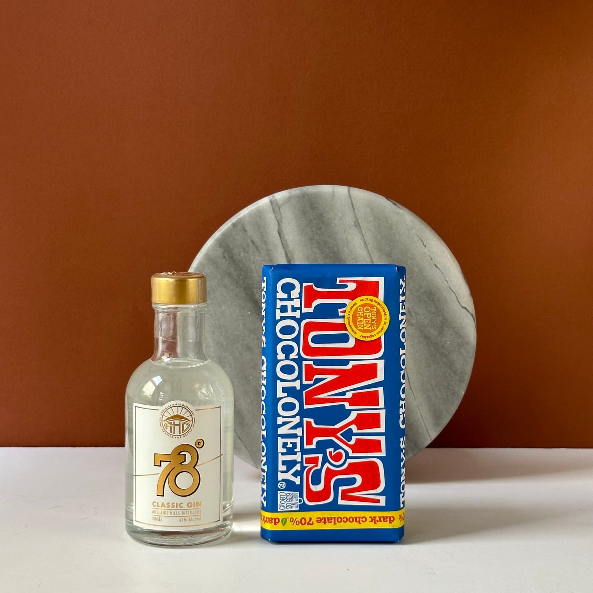 A product display featuring a small bottle of 78 Degrees Classic Gin next to a large bar of Tony's Chocolonely Dark Chocolate 70%. The items are set against a minimalist backdrop with a warm brown background and a round marble accent behind them