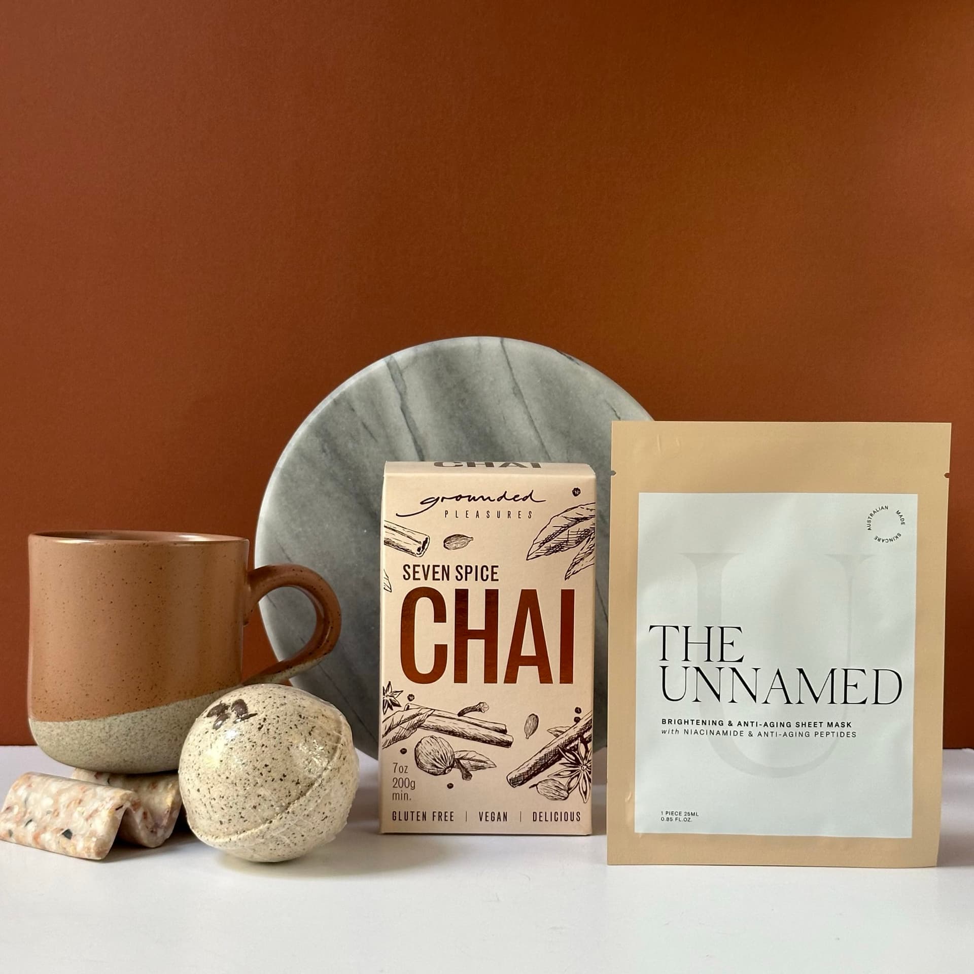 A collection of self-care items arranged on a white surface against a warm brown background. The set includes a Robert Gordon ceramic mug, a box of Grounded Pleasures Seven Spice Chai, a packet of The Unnamed brightening sheet mask, a round marble plate, a Mylk and Butter bath bomb,