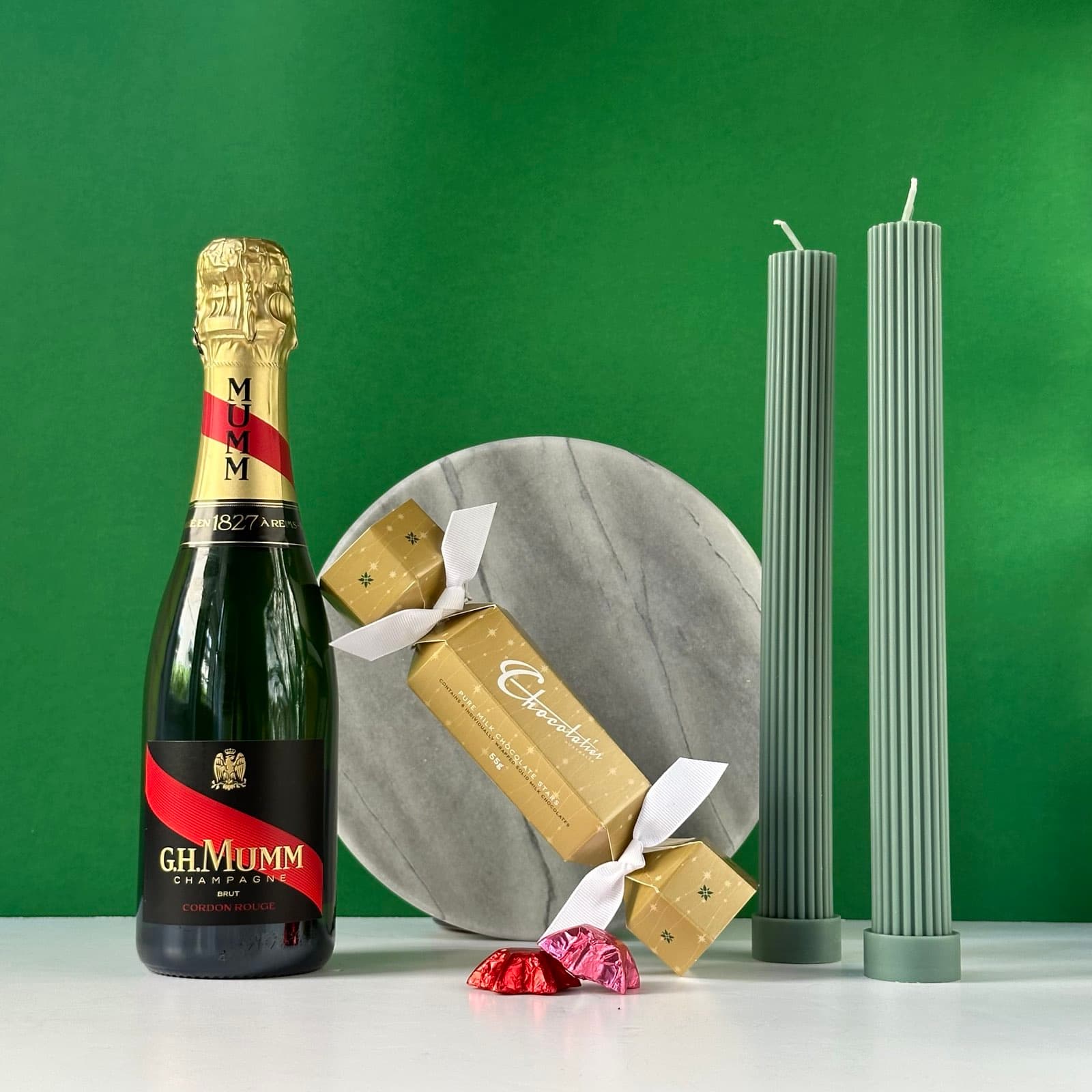 A luxury gift set arranged on a white surface against a green background. The set includes: A bottle of G.H. Mumm Cordon Rouge Champagne A gold Crackalackin bon bon (Christmas cracker) on a round marble serving plate Two tall, sage green pillar candles (Black Blaze brand) Two small red foil-wrapped chocolates or candies The items are artfully arranged to showcase an elegant and festive gift bundle, perfect for celebrations or special occasions.