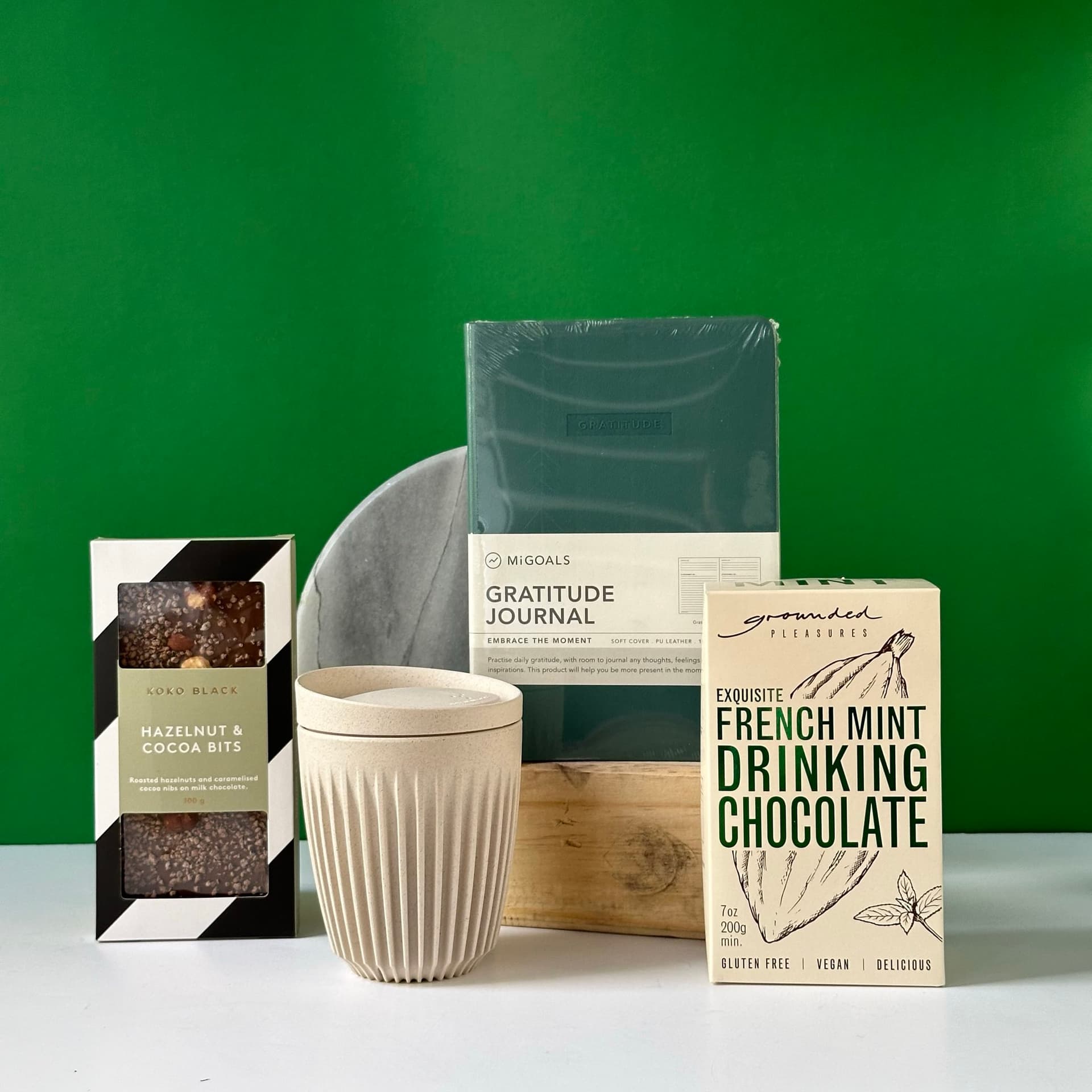 A collection of self-care items arranged on a white surface against a green background. The set includes a Mi Goals Gratitude Journal, a box of Koko Black Hazelnut & Cocoa Bits, a box of Grounded Pleasures French Mint Drinking Chocolate, a cream-colored ribbed ceramic mug, and a partial view of a round marble plate.