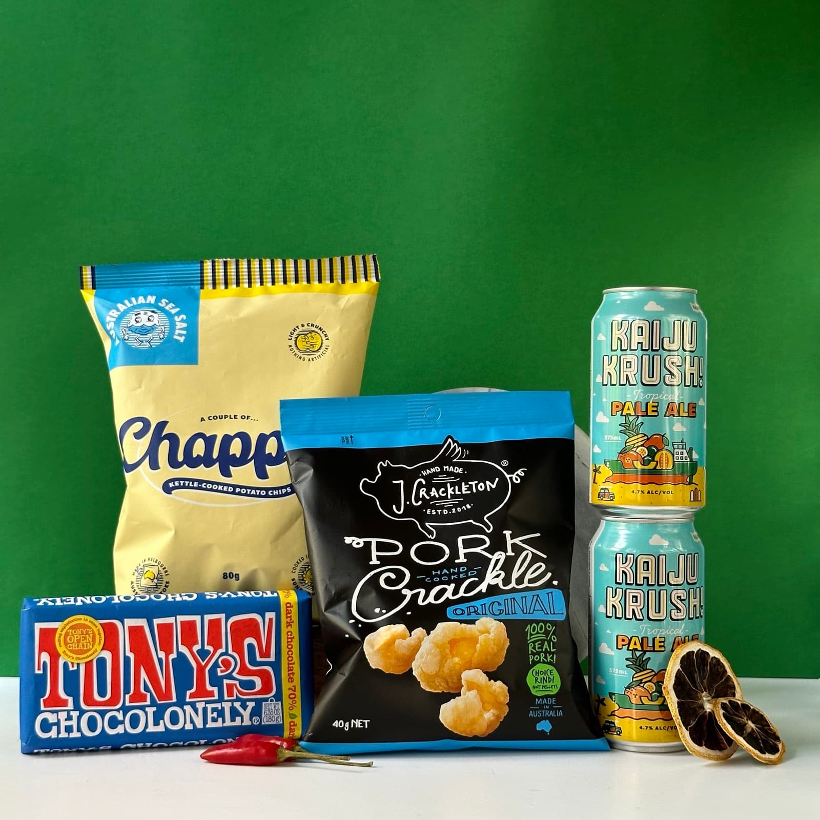 image.png PM Write an ALT text for website image ALT text: A collection of snacks and beverages on a white surface against a green background. The items include: A yellow bag of Chappy kettle-cooked potato chips A black bag of J. Crackleton's Pork Crackle Two cans of Kaiju Krush Tropical Pale Ale A blue box of Tony's Chocolonely chocolate A small red chili pepper A dried citrus slice The arrangement showcases a variety of Australian snack and drink options, presenting a colorful and diverse selection of treats.