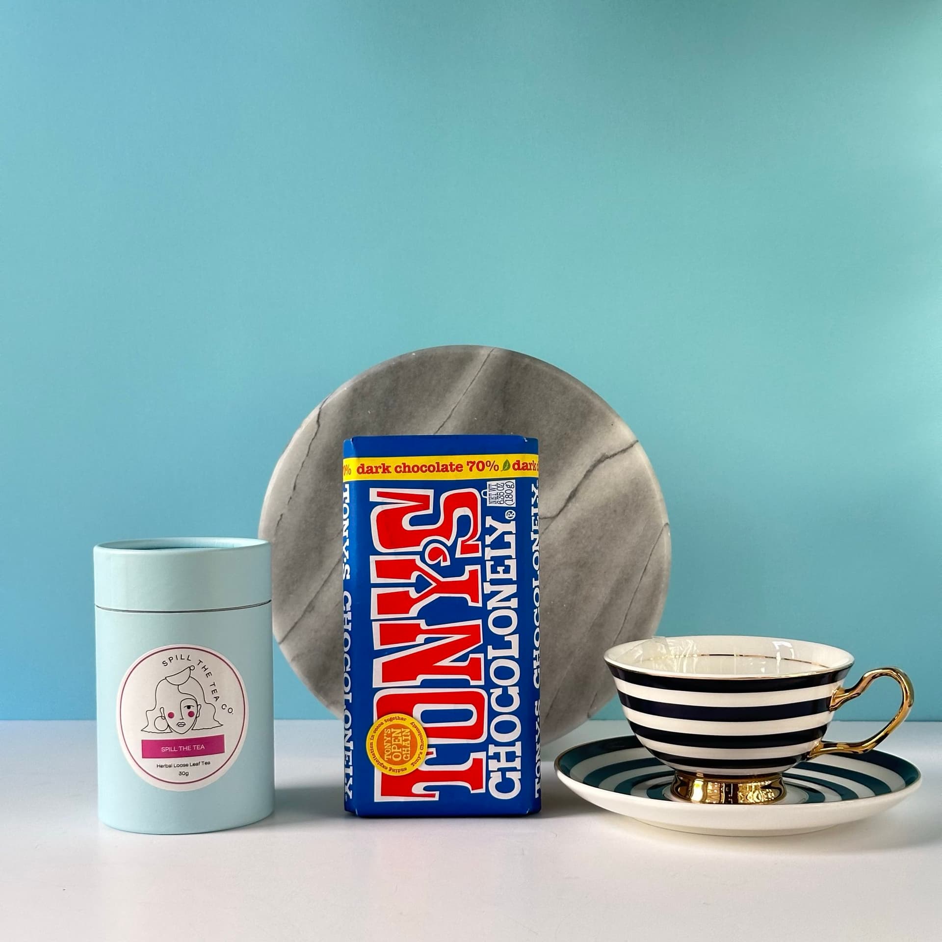 A gift set arranged on a white surface against a light blue background. The set includes a Tony's Chocolonely chocolate bar resting in a Lyndal T brand teacup with black and white stripes and a gold handle, a cylindrical container of Spill the Tea loose leaf tea, a round marble plate in the background, and a small wooden block. The teacup is placed on a matching striped saucer.