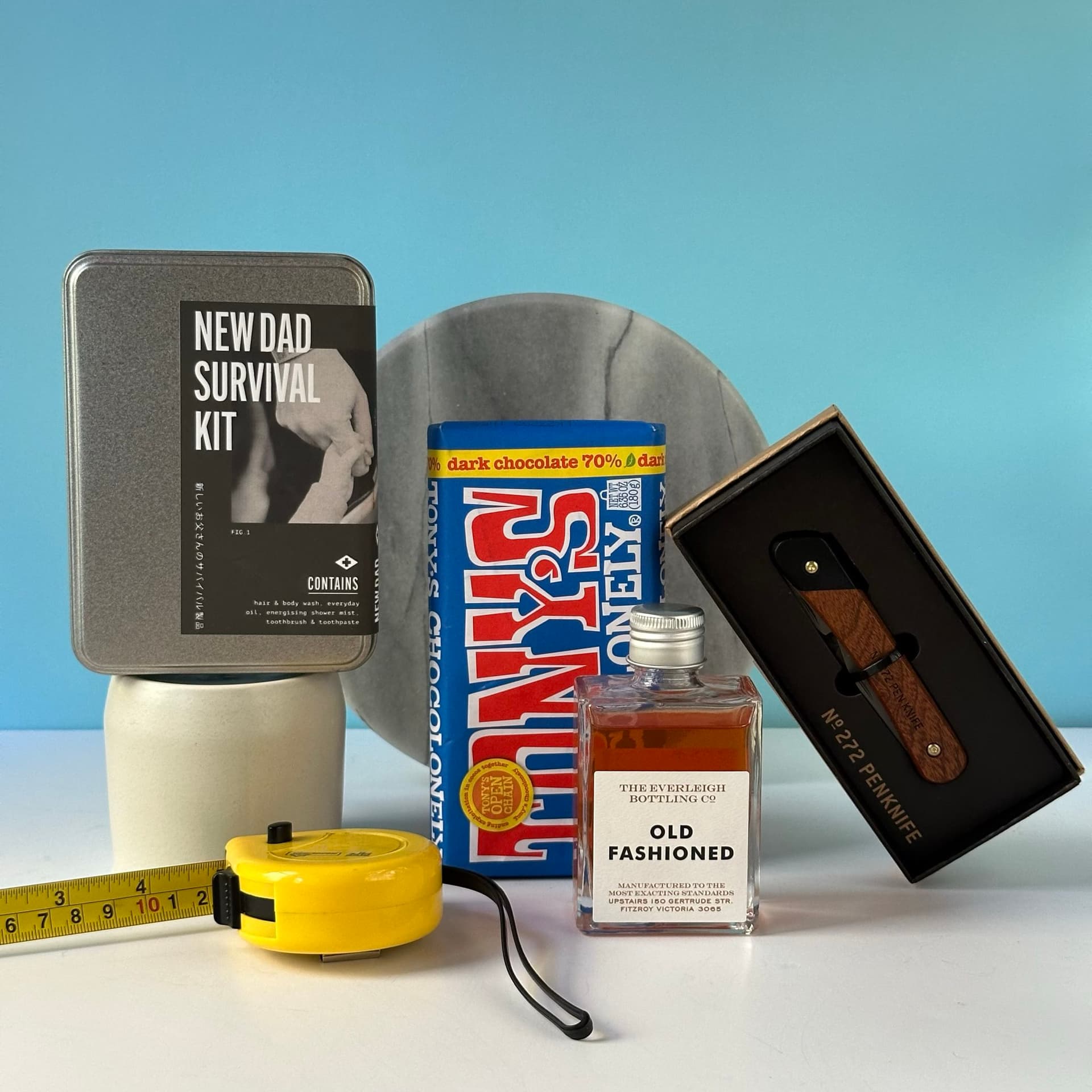 The "New Dad Kit" gift bundle arranged on a white surface against a light blue background. The set includes a metal tin labeled "New Dad Survival Kit", a Tony's Chocolonely dark chocolate bar, a small bottle of Old Fashioned cocktail from The Everleigh Bottling Co., a wooden-handled pocket knife in a box, a yellow tape measure, a round marble plate, and a white ceramic mug partially visible. These items are artfully arranged to create a thoughtful and practical gift set for new fathers.