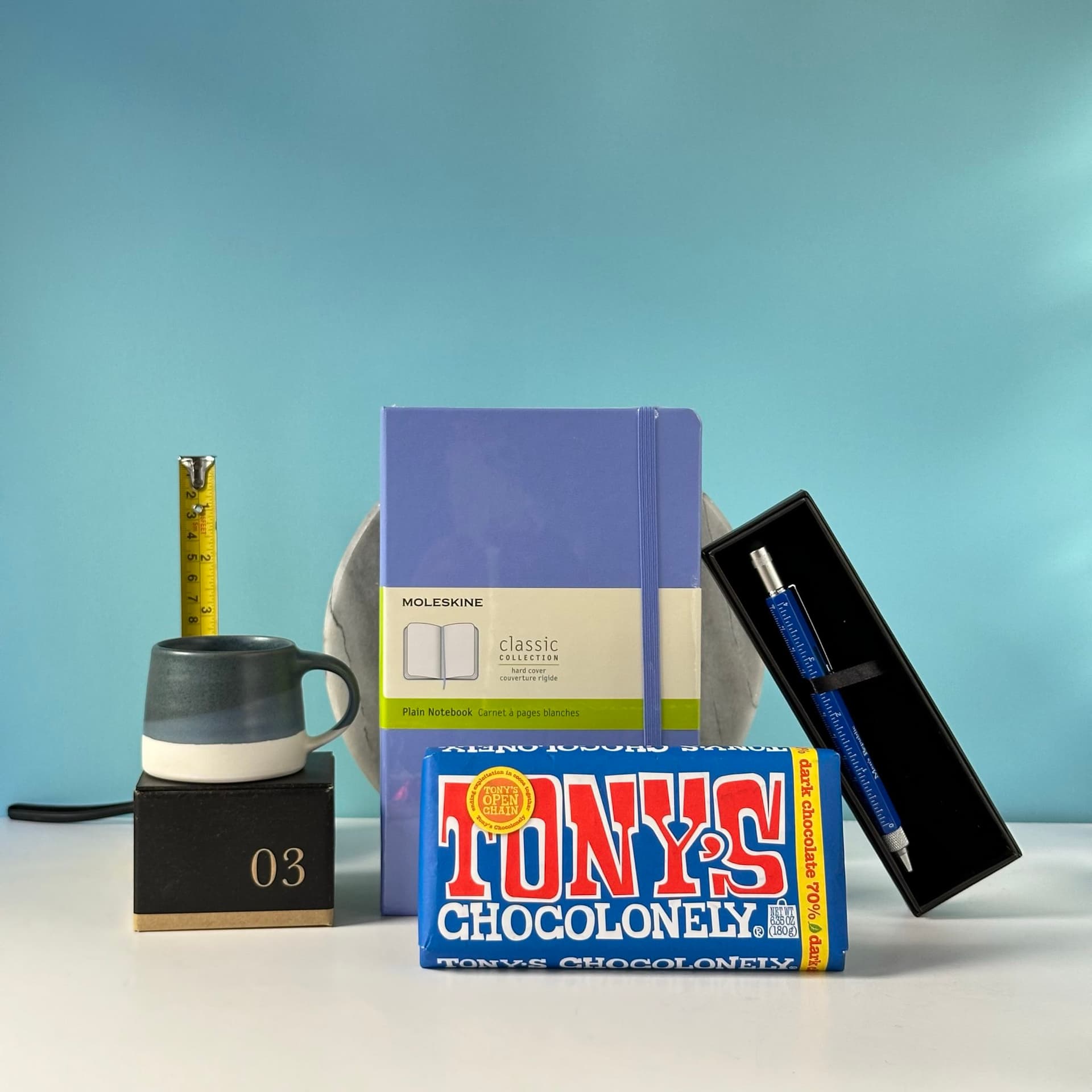 The Overthinker" gift bundle arranged on a white surface against a light teal background. The set includes a purple Moleskine notebook for endless thoughts, a Tony's Chocolonely chocolate bar for comfort, a ceramic mug for late-night pondering sessions, a yellow tape measure symbolizing the need to quantify everything, a sleek blue pen in a case for jotting down ideas, and a black cube marked "03". A round marble plate partially visible in the background adds a touch of sophistication to this thoughtfully curated collection for the chronic thinker.