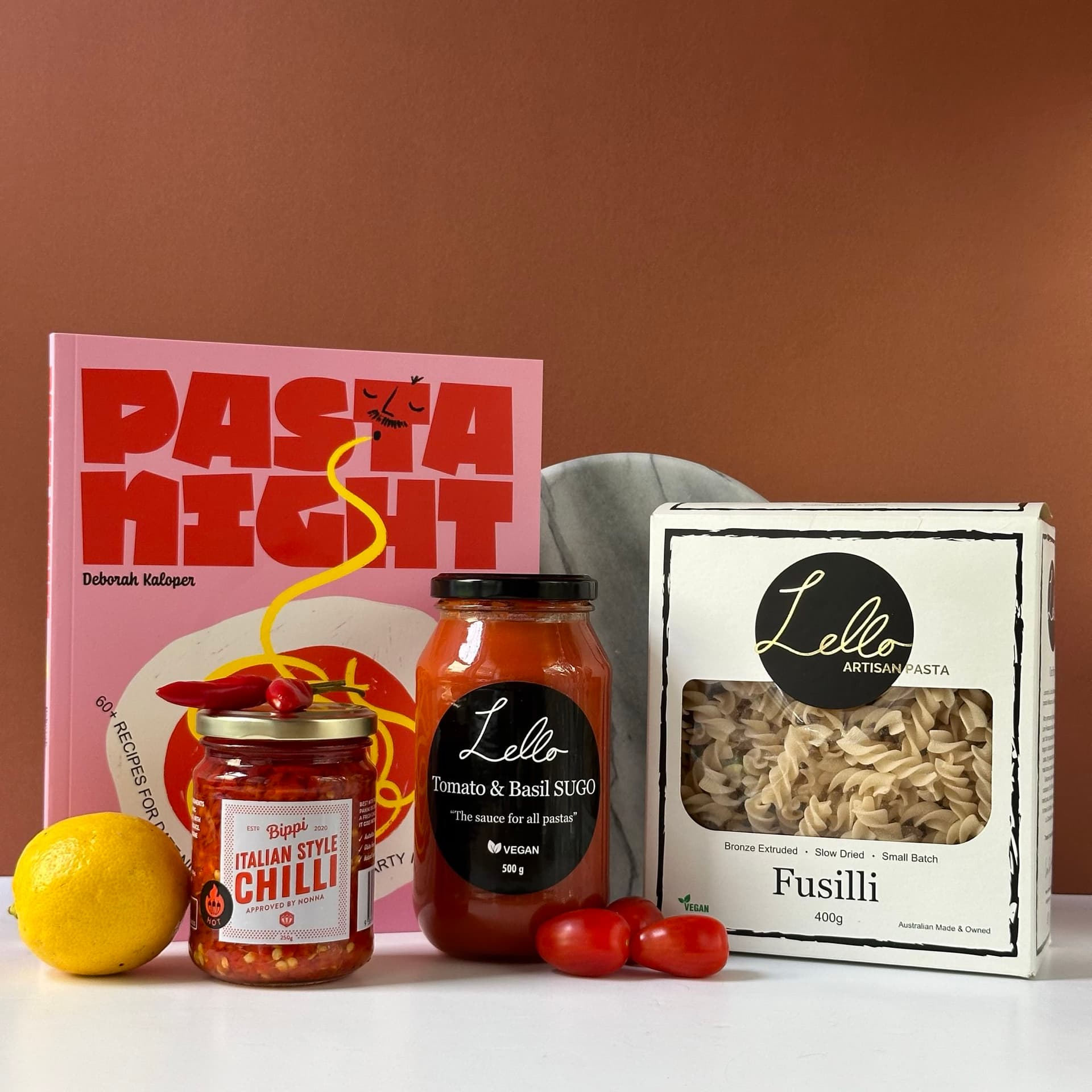 own background. The items include a pink cookbook titled "Pasta Night" by Deborah Kaloper, a jar of Bippi Italian Style Chilli, a jar of Lello Tomato & Basil Sugo, a box of Lello Fusilli pasta, fresh cherry tomatoes, and a lemon. A round marble plate is partially visible in the background.