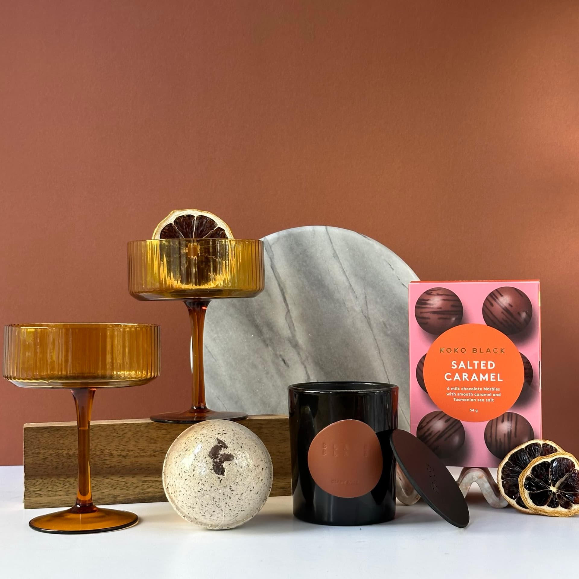 A luxurious gift set arranged on a white surface against a warm brown background. The set includes two amber-colored coupe glasses, a black scented candle, a speckled bath bomb, a box of Koko Black Salted Caramel Marbles, a round marble plate, and decorative dried citrus slices. The items are elegantly displayed, creating an atmosphere of sophistication and indulgence.