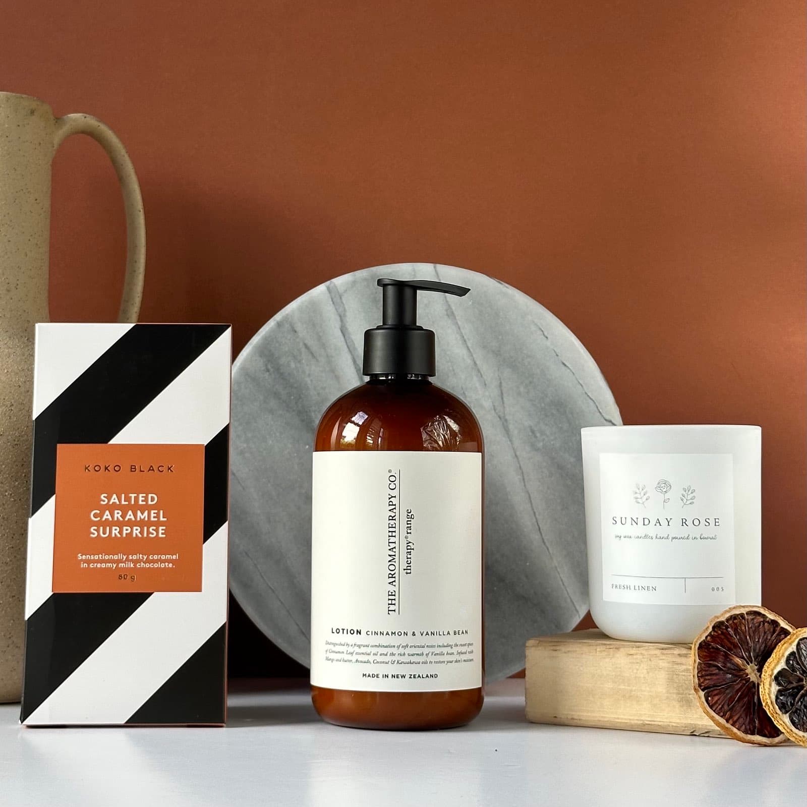 ALT text: The "Thank You" bundle featuring a collection of premium self-care products against a warm terracotta background. The items include: 1. A Koko Black "Salted Caramel Surprise" chocolate box with striking black and white diagonal stripes 2. A large amber glass bottle of "The Aromatherapy Co." lotion in Cinnamon & Vanilla Bean scent 3. A white "Sunday Rose" scented candle in a minimalist container 4. A round marble serving plate or coaster 5. A partial view of a beige ceramic mug 6. Dried citrus slices for decoration The arrangement is artfully composed on a light surface, showcasing a luxurious and cohesive gift set with warm, inviting tones and textures.