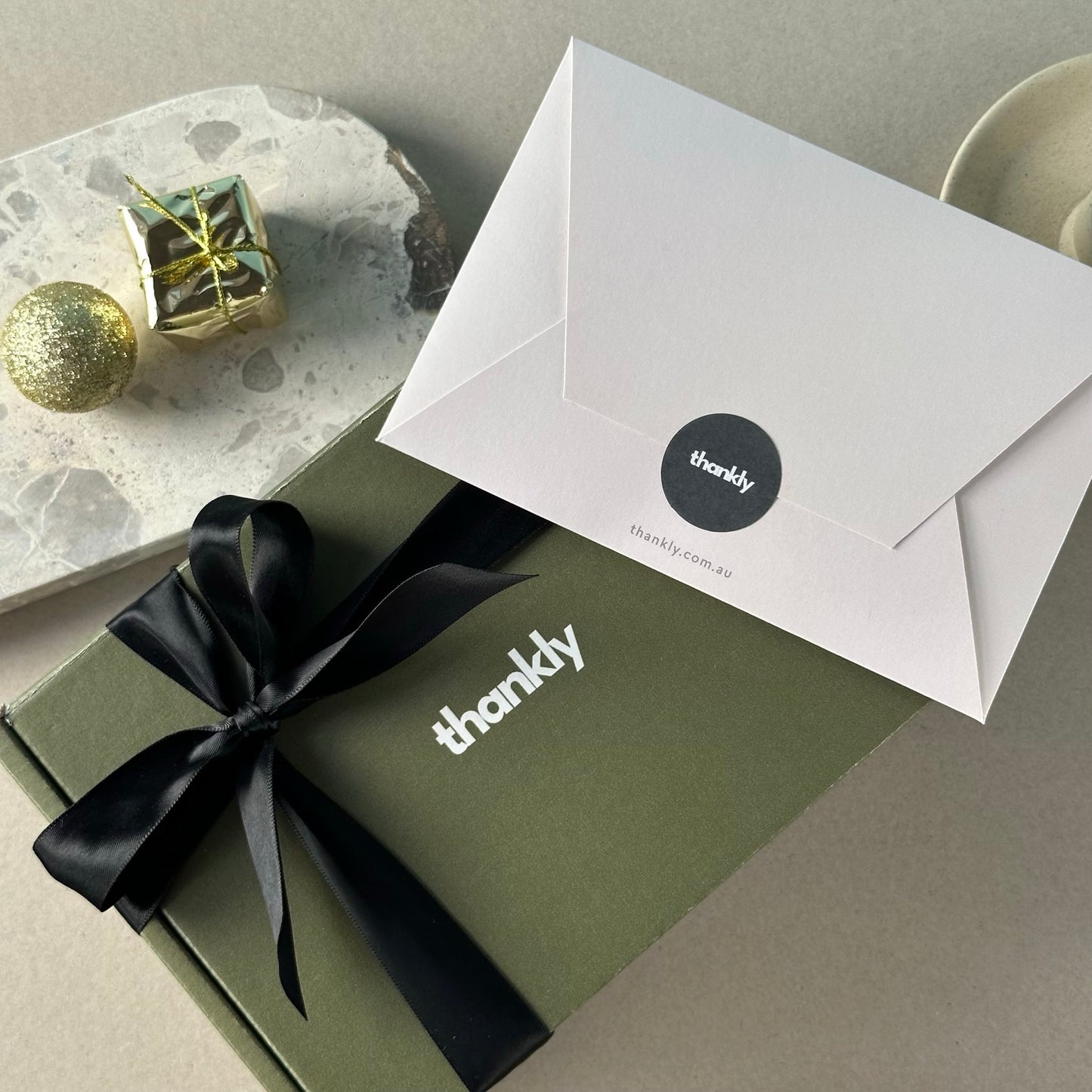 Green gift box with black ribbon and 'thankly' branding, accompanied by a white envelope on a neutral background.